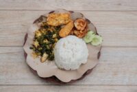 Sepiring nasi kuning pulen disajikan dengan lauk-pauk komplit seperti ayam goreng, telur balado, sambal goreng kentang, dan irisan timun segar, menggambarkan kelezatan serta kekayaan kuliner Nusantara. Hidangan ini memancarkan warna kuning cerah yang menggoda selera, siap memanjakan setiap penikmatnya.
