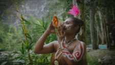 Seorang belian dari Suku Dayak Benuaq dengan pakaian adat lengkap sedang melakukan ritual penyembuhan, memperlihatkan kekayaan budaya dan spiritualitas yang mendalam di pedalaman Kalimantan. Gerakan tubuh dan ekspresi wajahnya memancarkan energi yang kuat, menjadi pusat perhatian dalam upacara adat yang penuh makna.