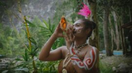 Seorang belian dari Suku Dayak Benuaq dengan pakaian adat lengkap sedang melakukan ritual penyembuhan, memperlihatkan kekayaan budaya dan spiritualitas yang mendalam di pedalaman Kalimantan. Gerakan tubuh dan ekspresi wajahnya memancarkan energi yang kuat, menjadi pusat perhatian dalam upacara adat yang penuh makna.