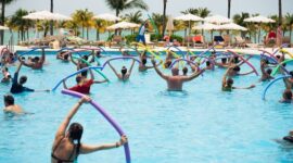 Sekelompok wanita lanjut usia tersenyum ceria saat melakukan gerakan aqua aerobik di dalam kolam renang, menunjukkan betapa olahraga air dapat dinikmati oleh semua kalangan usia. Air memberikan dukungan yang lembut sehingga latihan kebugaran terasa lebih ringan dan menyenangkan.